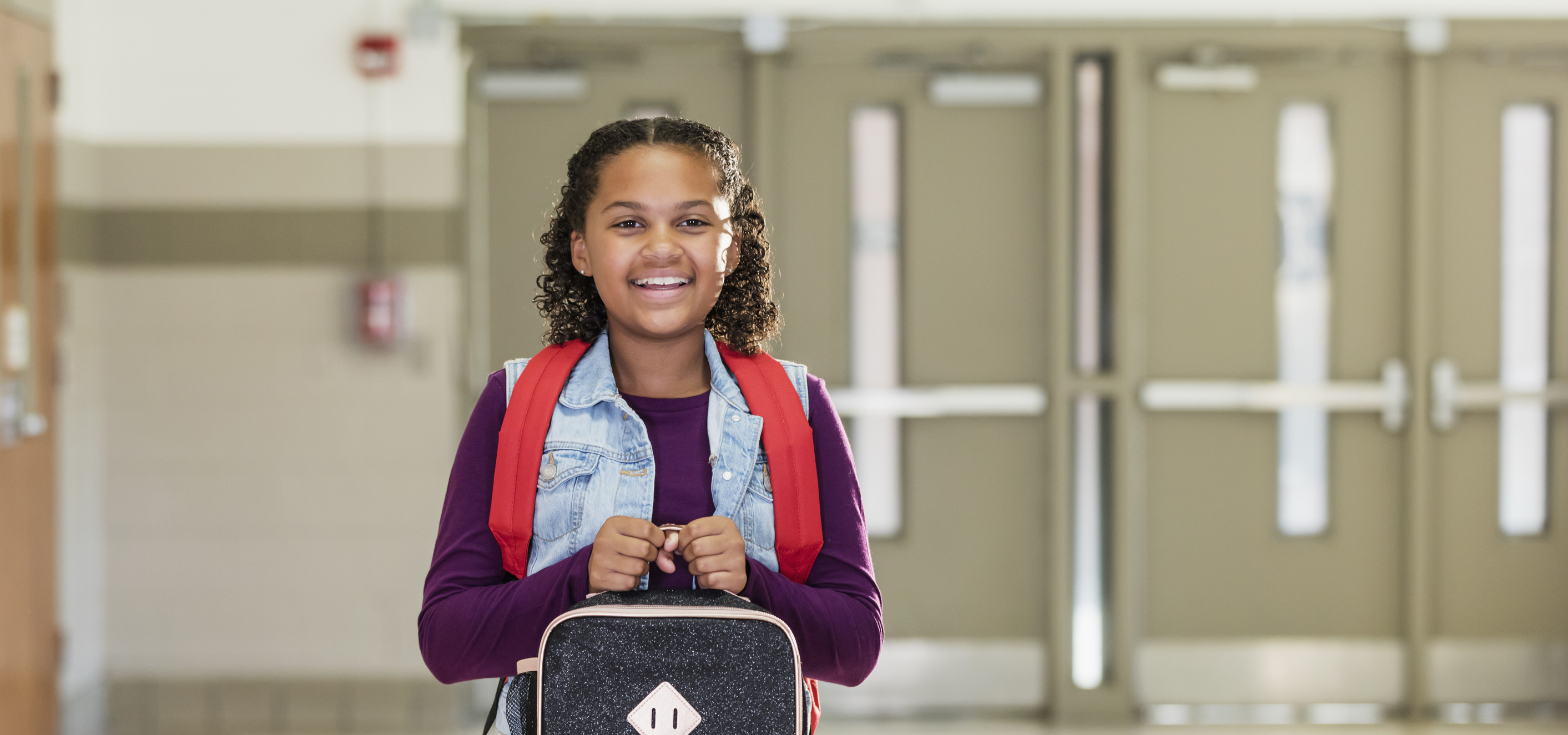 A 10 year old girl is standing ans smiling in the hallway of her elementary school carrying a backpack and lunch box looking very happy, with out of focus emergency doors in the background equipped with panic devices, door closers, and kick plates.